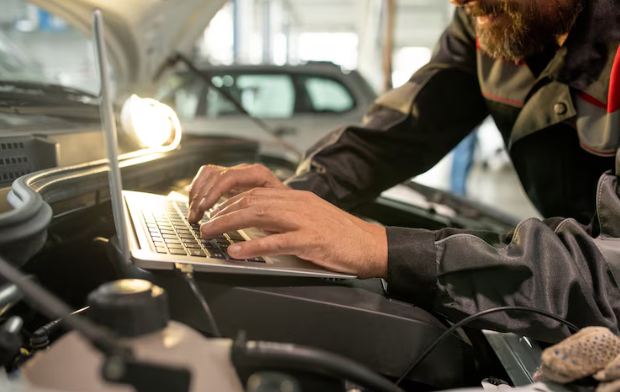 Hands of middle aged male worker of car maintenance service bending over open engine compartment in workshop while using laptop